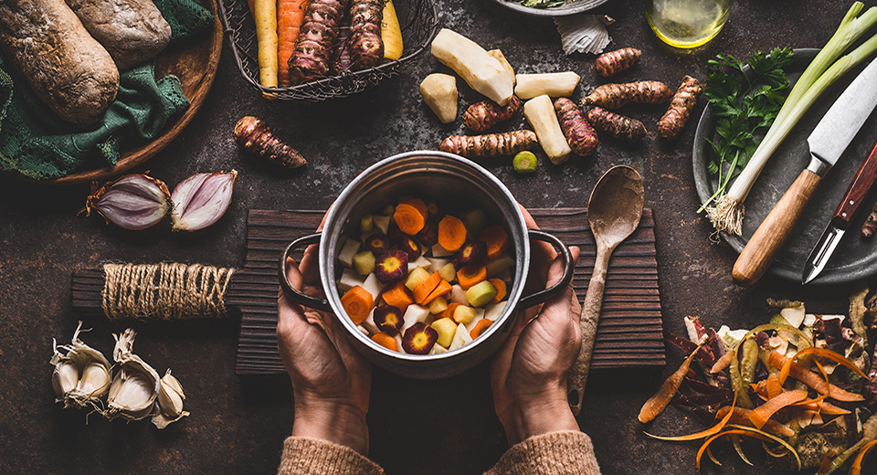 Female woman hands holding pan with diced colorful vegetables on dark rustic kitchen table with vegetarian cooking ingredients and tools. Healthy and clean seasonal food cooking and eating concept